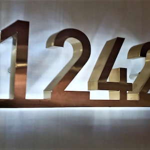 Könnte beinhalten: Ein goldfarbenes Metallschild mit der Nummer 1242 darauf. Das Schild wird von hinten beleuchtet, wodurch ein helles Leuchten um die Zahlen entsteht.