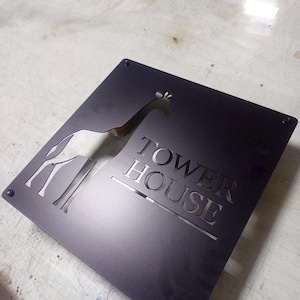 May include: Black metal sign with a giraffe silhouette and the words "TOWER HOUSE" cut out. The sign is square with rounded corners and has four screw holes. The giraffe is facing left.