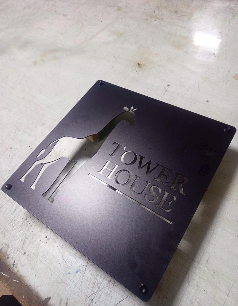 May include: Black metal sign with a giraffe silhouette and the words "TOWER HOUSE" cut out. The sign is square with rounded corners and has four screw holes. The giraffe is facing left.