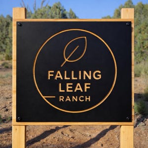 Op de afbeelding: Een rechthoekig zwart metalen bord gemonteerd op een lichtgekleurd houten frame. Het bord heeft een cirkelvormig uitgesneden ontwerp met een bladpictogram en de tekst FALLING LEAF RANCH in het midden gegraveerd. Vier zwarte bouten.