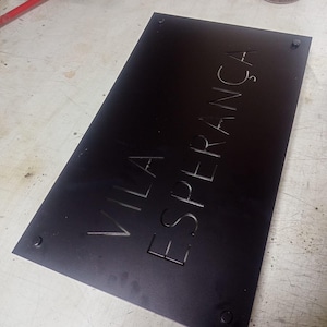 May include: A black rectangular sign with the words "VILA ESPERAN&Ccedil;A" engraved in a sans-serif font. The sign is secured with four screws at each corner. The sign is likely for a business or residence.