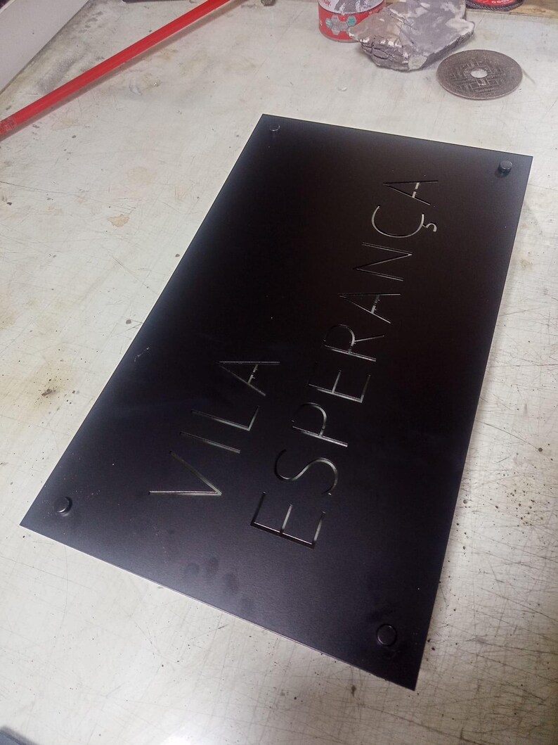 May include: A black rectangular sign with the words "VILA ESPERAN&Ccedil;A" engraved in a sans-serif font. The sign is secured with four screws at each corner. The sign is likely for a business or residence.