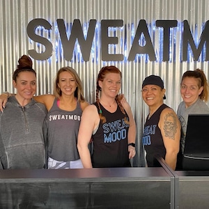 May include: Five people smiling in front of a corrugated metal wall with the word "SWEATMOOD" in large, illuminated letters. The people are wearing athletic apparel, including tank tops and a zip-up hoodie. The tank tops have the words "SWEATMOOD" printed on them.