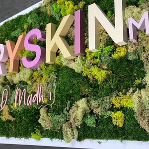 May include: A wall-mounted sign featuring three-dimensional letters spelling EVERY SKIN MD in shades of pink, beige, and tan, set against a textured background of preserved green moss and lichen, with a clear acrylic sign reading by D Madh below.