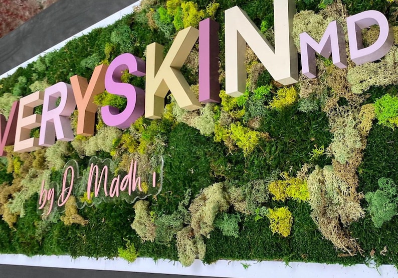 May include: A wall-mounted sign featuring three-dimensional letters spelling EVERY SKIN MD in shades of pink, beige, and tan, set against a textured background of preserved green moss and lichen, with a clear acrylic sign reading by D Madh below.
