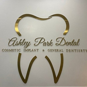 以下が含まれることがあります： 「Ashley Park Dental」という文字と、「Cosmetic Implant & General Dentistry」という文字が書かれた、金色の歯の形をした看板です。