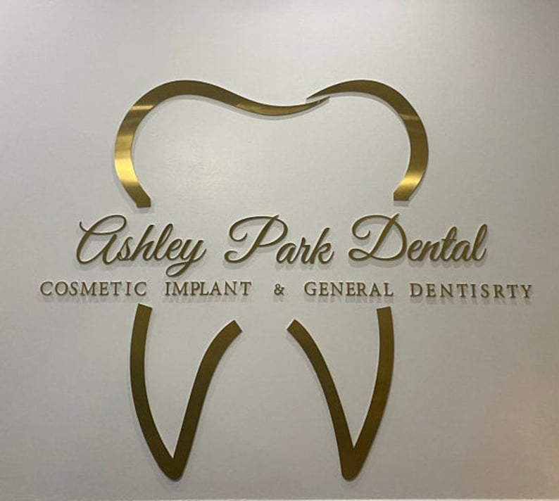 以下が含まれることがあります： 「Ashley Park Dental」という文字と、「Cosmetic Implant & General Dentistry」という文字が書かれた、金色の歯の形をした看板です。