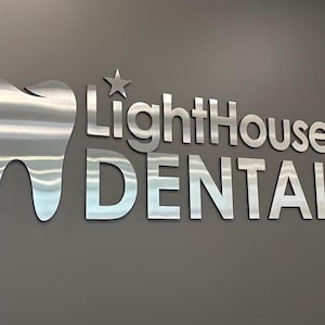 May include: A three-dimensional wall sign featuring a brushed silver metal tooth icon next to a five-pointed star and the text LightHouse DENTAL in a bold, sans-serif font. The metallic letters and icon are mounted against a solid gray wall.