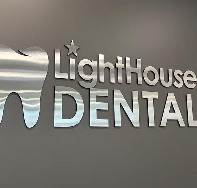 May include: A three-dimensional wall sign featuring a brushed silver metal tooth icon next to a five-pointed star and the text LightHouse DENTAL in a bold, sans-serif font. The metallic letters and icon are mounted against a solid gray wall.