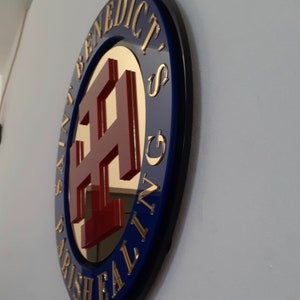 May include: A circular wall sign featuring a dark blue border with gold lettering that reads SAINT BENEDICT'S PARISH EALING. The center contains a reflective gold surface with a raised, deep red cross symbol mounted in the middle of the plaque.