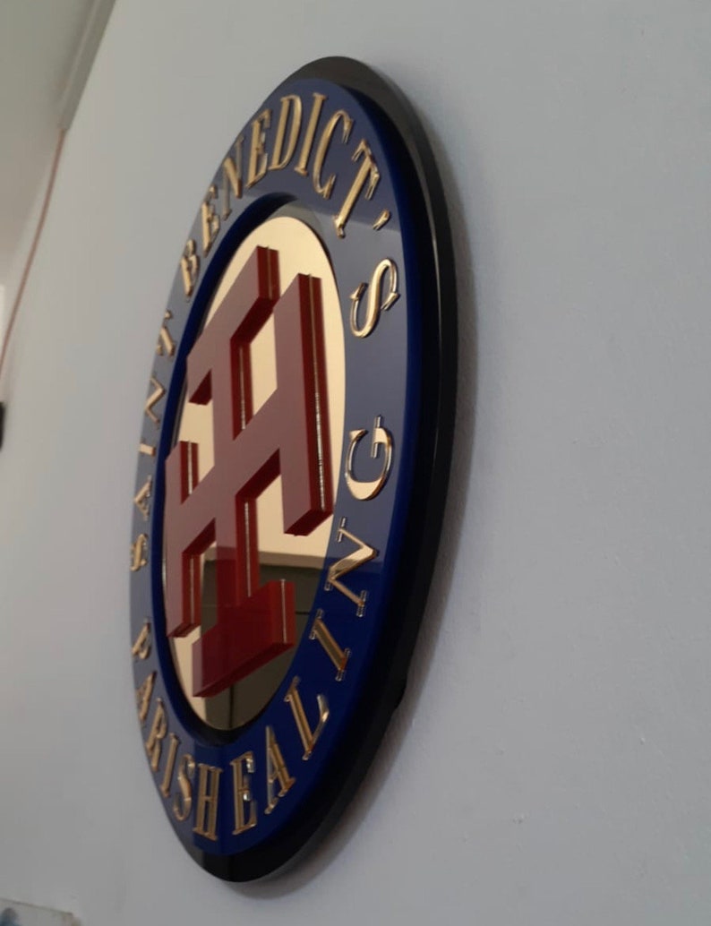 May include: A circular wall sign featuring a dark blue border with gold lettering that reads SAINT BENEDICT'S PARISH EALING. The center contains a reflective gold surface with a raised, deep red cross symbol mounted in the middle of the plaque.