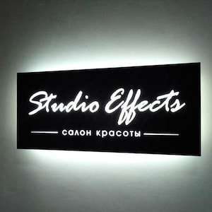 May include: A black rectangular sign with white lettering that reads "Studio Effects salon красоты". The sign is illuminated with white light.