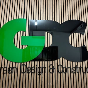 May include: A wall-mounted sign featuring the large letters GDC in a combination of glossy green and black finishes. Below the letters, the text Green Design & Construct is displayed in a clean, black, sans-serif font on a slatted wood wall.