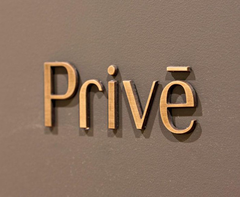 K&ouml;nnte beinhalten: Nahaufnahme des Wortes "Priv&eacute;" in goldfarbenen, erhabenen Buchstaben. Die Buchstaben heben sich von einem ged&auml;mpften, dunkelgrauen Hintergrund ab. Das Schild wird wahrscheinlich f&uuml;r einen privaten Bereich oder eine Einrichtung verwendet.