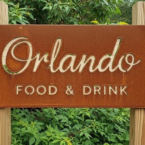 May include: A weathered, rectangular sign with the word "Orlando" cut out in a decorative font, above the words "FOOD & DRINK". The sign is mounted on two wooden posts and surrounded by greenery.