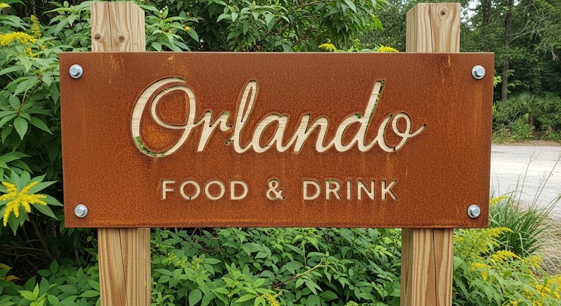 May include: A weathered, rectangular sign with the word "Orlando" cut out in a decorative font, above the words "FOOD & DRINK". The sign is mounted on two wooden posts and surrounded by greenery.