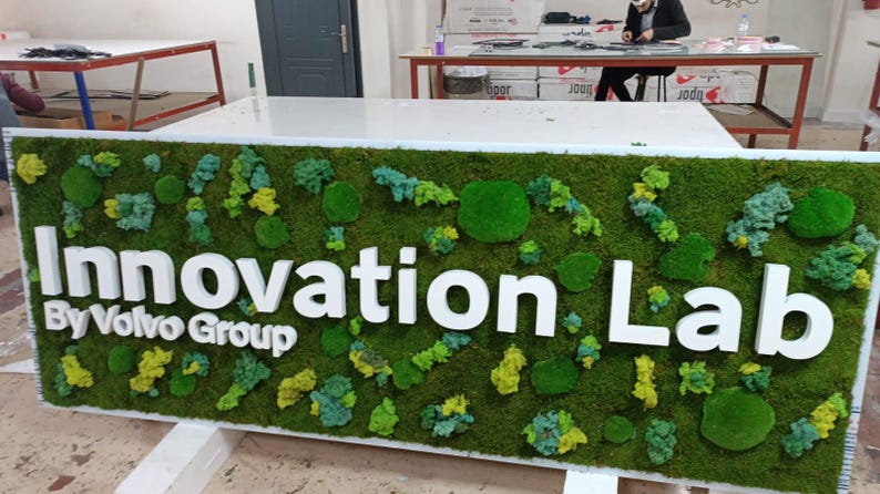 May include: A rectangular sign featuring a vibrant green moss background with various shades of preserved moss clusters. The white 3D lettering reads Innovation Lab By Volvo Group, mounted prominently against the textured natural moss surface.
