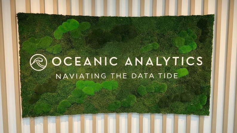 May include: A rectangular wall sign featuring a dense, textured green moss background. The sign displays the white text "OCEANIC ANALYTICS" above the phrase "NAVIATING THE DATA TIDE" and a circular icon containing a stylized wave graphic.