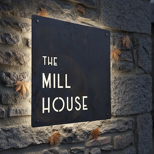 May include: A black rectangular sign with the words "THE MILL HOUSE" in white, mounted on a stone wall. The sign is illuminated by a light, and several brown leaves are scattered around it.