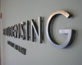 May include: Metal letters spelling out "REUDENISING" and "ATTORNEYS AT LAW" are mounted on a light gray wall. The letters are in a sans-serif font and have a brushed metal finish. The letter "G" is particularly large.