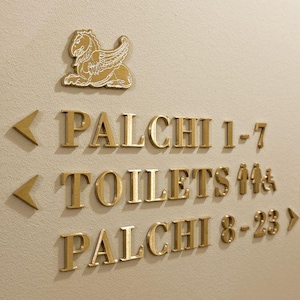 K&ouml;nnte beinhalten: Goldfarbene Wegweiser an einer beigefarbenen Wand. Das Schild enth&auml;lt die W&ouml;rter "PALCHI 1-7", "TOILETTEN" und "PALCHI 8-23" sowie ein gefl&uuml;geltes Pferdeemblem. Pfeile zeigen die Richtung an.