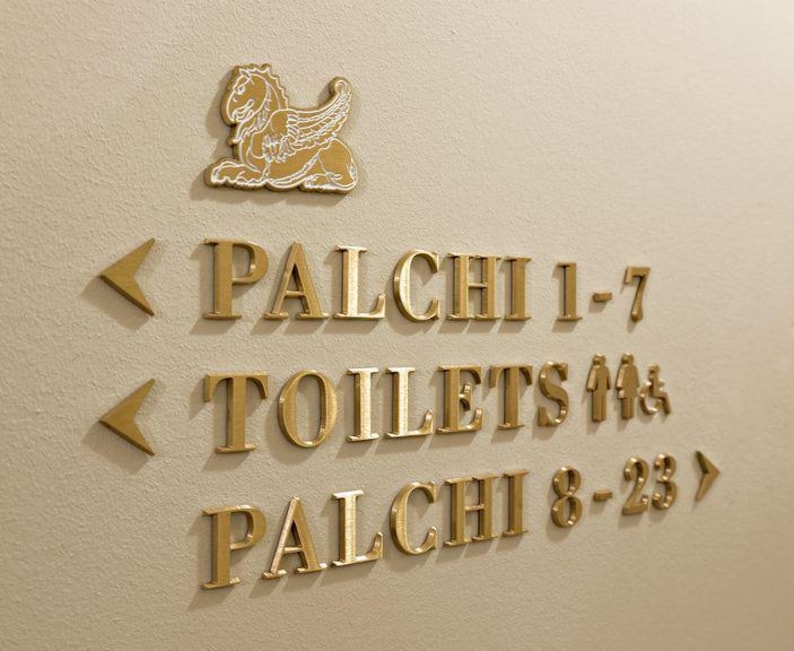 K&ouml;nnte beinhalten: Goldfarbene Wegweiser an einer beigefarbenen Wand. Das Schild enth&auml;lt die W&ouml;rter "PALCHI 1-7", "TOILETTEN" und "PALCHI 8-23" sowie ein gefl&uuml;geltes Pferdeemblem. Pfeile zeigen die Richtung an.