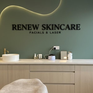 May include: A sage green wall features the black text RENEW SKINCARE FACIALS & LASER with a curved LED light strip above. Below, a white countertop holds a round sink, a gold faucet, various skincare bottles, a wax warmer, and a small mirror.