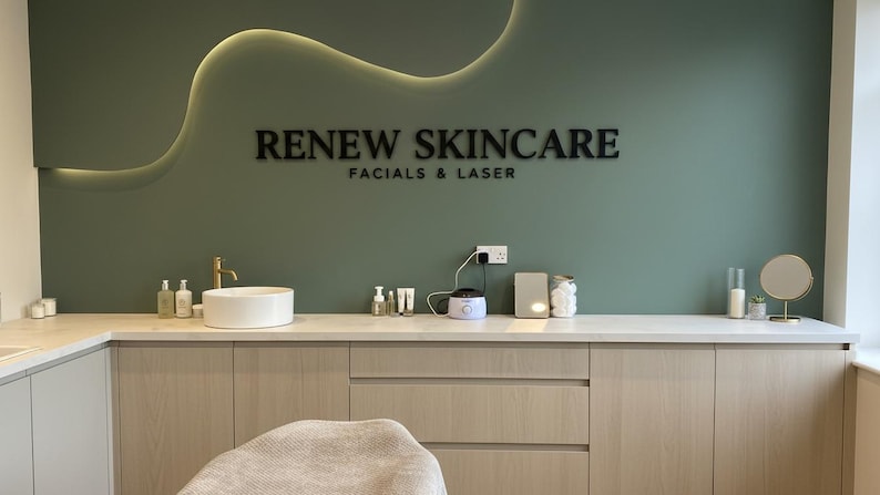 May include: A sage green wall features the black text RENEW SKINCARE FACIALS & LASER with a curved LED light strip above. Below, a white countertop holds a round sink, a gold faucet, various skincare bottles, a wax warmer, and a small mirror.