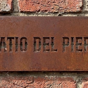 May include: A rectangular, weathered metal sign with the words "PATIO DEL PIERCE" cut out. The sign is mounted on a red brick wall with four screws. The sign has a rusty, brown color.