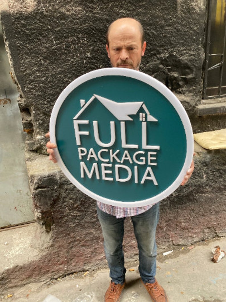 May include: A circular sign with a teal background and a white border. The sign features a white house icon above the white text "FULL PACKAGE MEDIA" in a sans-serif font. The design is three-dimensional with raised white lettering and graphics.