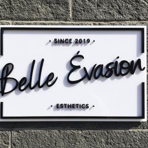 May include: A rectangular white sign with a black border featuring the black text Belle &Eacute;vasion in a cursive script. Above the text is the phrase SINCE 2019 and below is the word ESTHETICS, both in a sans-serif font with small black dots.