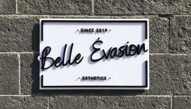May include: A rectangular white sign with a black border featuring the black text Belle &Eacute;vasion in a cursive script. Above the text is the phrase SINCE 2019 and below is the word ESTHETICS, both in a sans-serif font with small black dots.