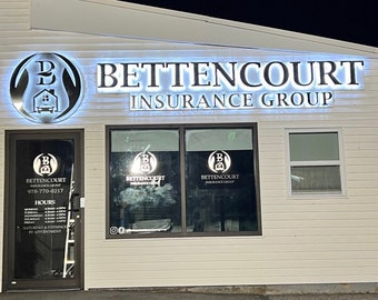 Könnte beinhalten: Außenansicht des Bürogebäudes der Bettencourt Insurance Group bei Nacht. Das Gebäude hat eine weiße Fassade mit beleuchteten Schildern. Das Logo zeigt ein stilisiertes "B" mit einem Haus- und Autosymbol. Die Öffnungszeiten sind an der Tür angegeben.