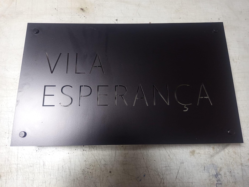 May include: A black metal sign with the words "VILA ESPERAN&Ccedil;A" cut out. The sign is rectangular and has four small, round fasteners in each corner. The text is in a sans-serif font.