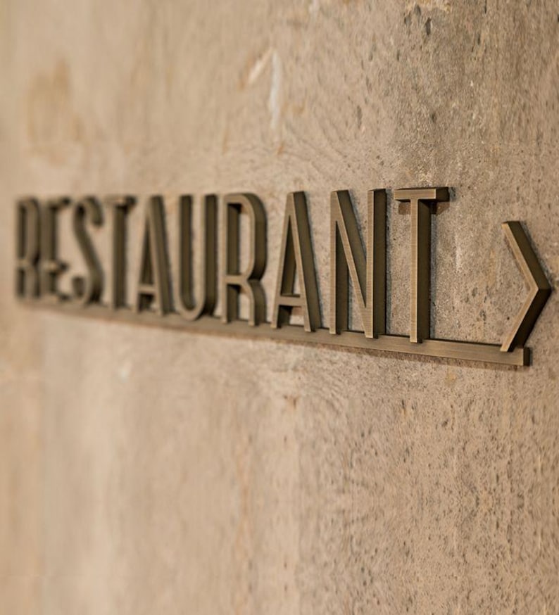K&ouml;nnte beinhalten: Ein bronzefarbenes Metallschild mit der Aufschrift "RESTAURANT" und einem Pfeil nach rechts. Das Schild ist an einer strukturierten, hellbraunen Wand angebracht.