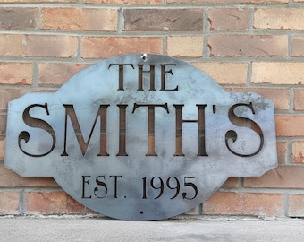 May include: A custom oval-shaped metal sign with a raw steel finish. The sign features laser-cut lettering that reads THE SMITH'S and EST. 1995. The design includes a mounting hole at the top and bottom center for installation on a flat surface.
