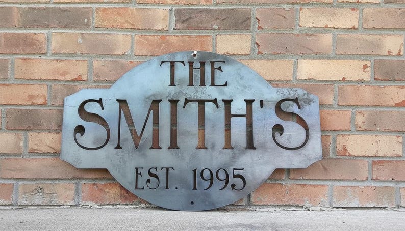 May include: A metal sign with the text "THE SMITH'S EST. 1995" cut out. The sign is a light gray color and is mounted on a brick wall. The sign has a decorative shape.