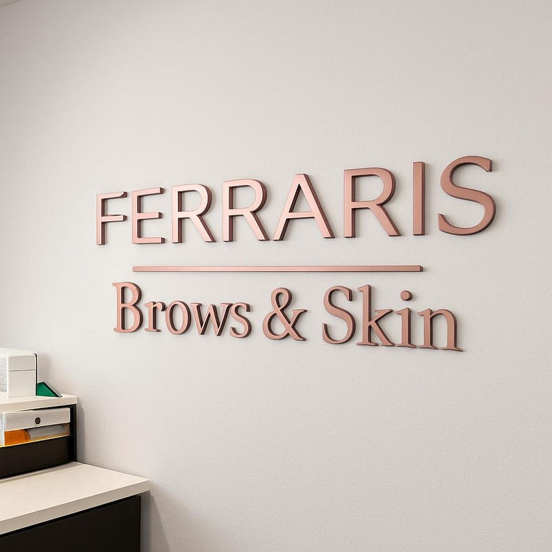 May include: A wall-mounted sign featuring three-dimensional rose gold lettering that reads FERRARIS above a horizontal line and the text Brows & Skin. The metallic letters are mounted on a plain, light-colored wall surface in a professional setting.
