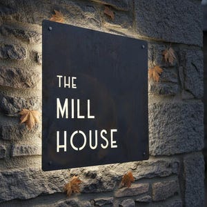 Op de afbeelding: Een vierkant, donker metalen bord met de tekst THE MILL HOUSE uitgesneden in een sjabloonlettertype. Het bord is bevestigd aan een ruwe stenen muur en heeft ge&iuml;ntegreerde achtergrondverlichting die de muur achter de metalen plaat verlicht.