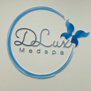 以下が含まれることがあります： 円の中に「DLux Medspa」という文字と、スタイリッシュな花のデザインが施された青と白のロゴです。