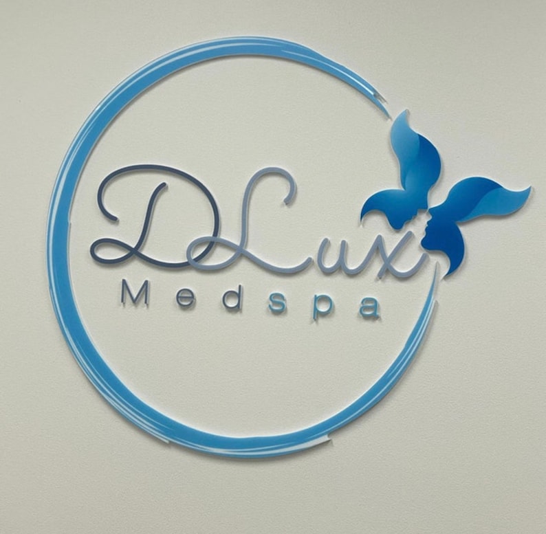 以下が含まれることがあります： 円の中に「DLux Medspa」という文字と、スタイリッシュな花のデザインが施された青と白のロゴです。