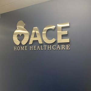 Puede incluir: Un letrero de metal dorado con la palabra "ACE" y la frase "HOME HEALTHCARE" en una pared gris.