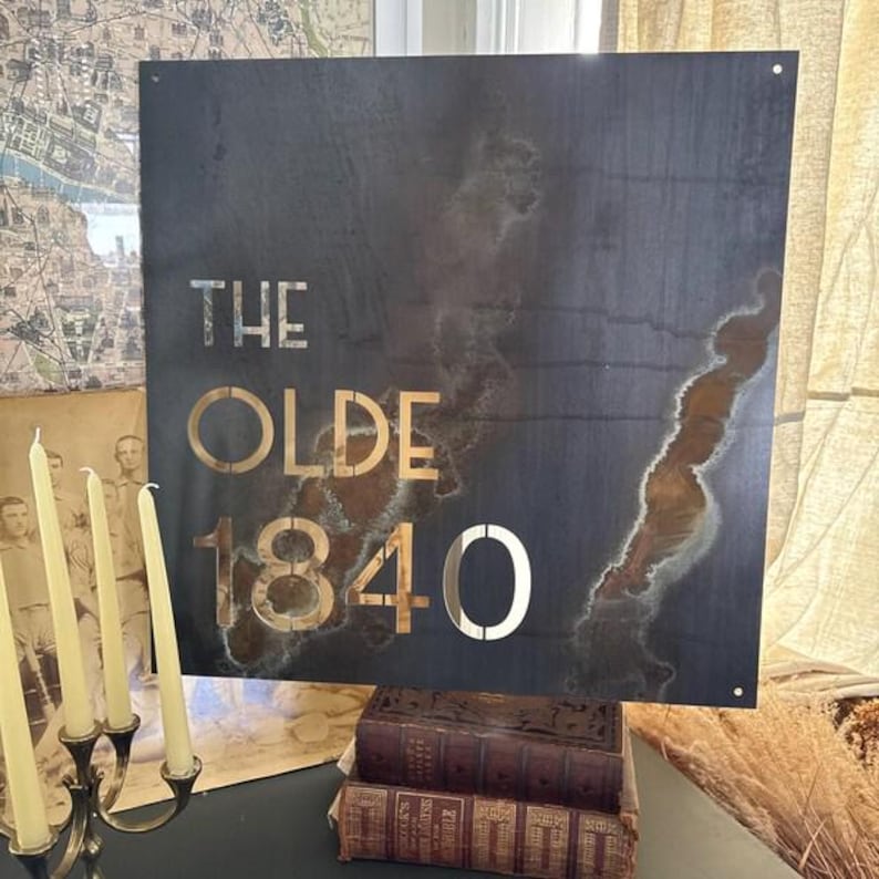 May include: A square metal sign with the words "THE OLDE 1840" cut out. The sign has a dark, weathered appearance with areas of rust-like coloration. The sign is displayed above a stack of old books.