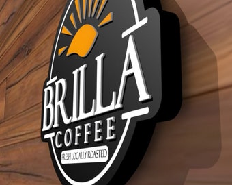 May include: A black, oval-shaped wall sign featuring the white text BRILLA COFFEE and the phrase FRESH LOCALLY ROASTED. The sign includes a stylized orange sun graphic at the top, with white border accents and a three-dimensional design.