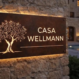 May include: A rectangular, dark, weathered metal sign with a cutout of an olive tree on the left side and the text "CASA WELLMANN" on the right. The sign is backlit with warm light, creating a glowing effect around the edges and the tree cutout.