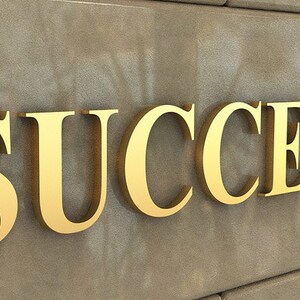 Puede incluir: La palabra "SUCCESS" en letras doradas sobre una pared de textura marr&oacute;n.