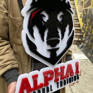 May include: A custom sign featuring a large, layered black and white wolf head logo mounted on a white acrylic backing. Below the wolf head, a separate white acrylic piece displays the red text ALPHA and black text PERSONAL TRAINING.