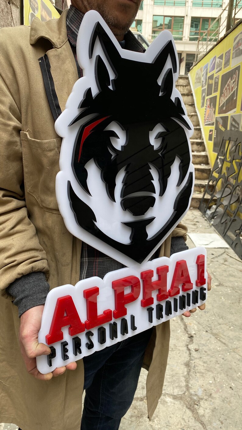 May include: A custom sign featuring a large, layered black and white wolf head logo mounted on a white acrylic backing. Below the wolf head, a separate white acrylic piece displays the red text ALPHA and black text PERSONAL TRAINING.