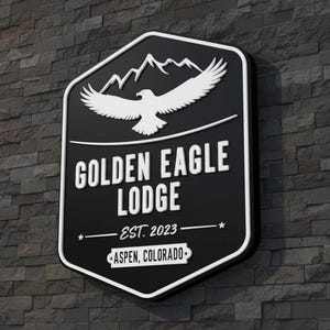 May include: A black, shield-shaped wall sign featuring a white graphic of a soaring eagle above mountain peaks. The text reads GOLDEN EAGLE LODGE, EST. 2023, and ASPEN, COLORADO. The sign has a thick white border and is mounted on a dark stone wall.
