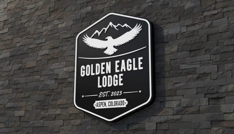 May include: A black, shield-shaped wall sign featuring a white graphic of a soaring eagle above mountain peaks. The text reads GOLDEN EAGLE LODGE, EST. 2023, and ASPEN, COLORADO. The sign has a thick white border and is mounted on a dark stone wall.